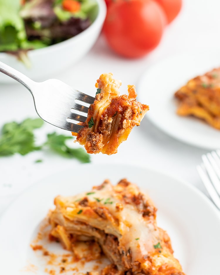 A bite of lasagna on a fork, above a piece of it on the plate.