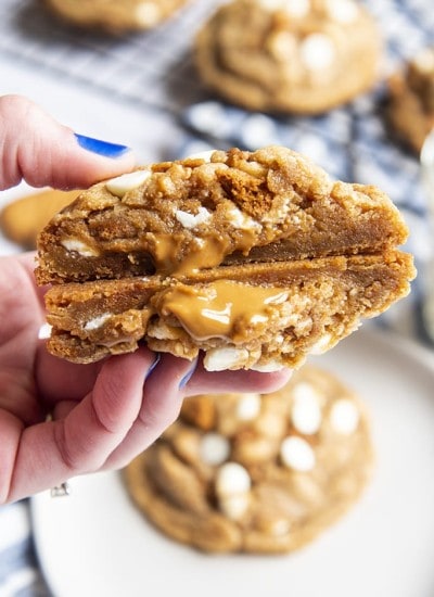 A hand holding a giant cookie that is cut in half showing the inside of the cookie. It looks slightly gooey, and there are white chocolate chips, and cookie butter spread oozing out.