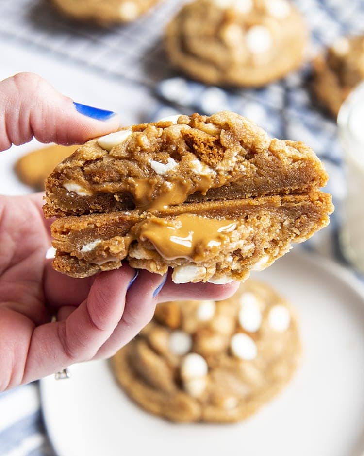 A hand holding a giant cookie that is cut in half showing the inside of the cookie. It looks slightly gooey, and there are white chocolate chips, and cookie butter spread oozing out.