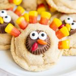 A close up shot of a peanut butter cookie decorated to look like a turkey with a peanut butter cup decorated with candy eyes, and chocolate covered candies for a beak, and candy corn at the top for feathers.