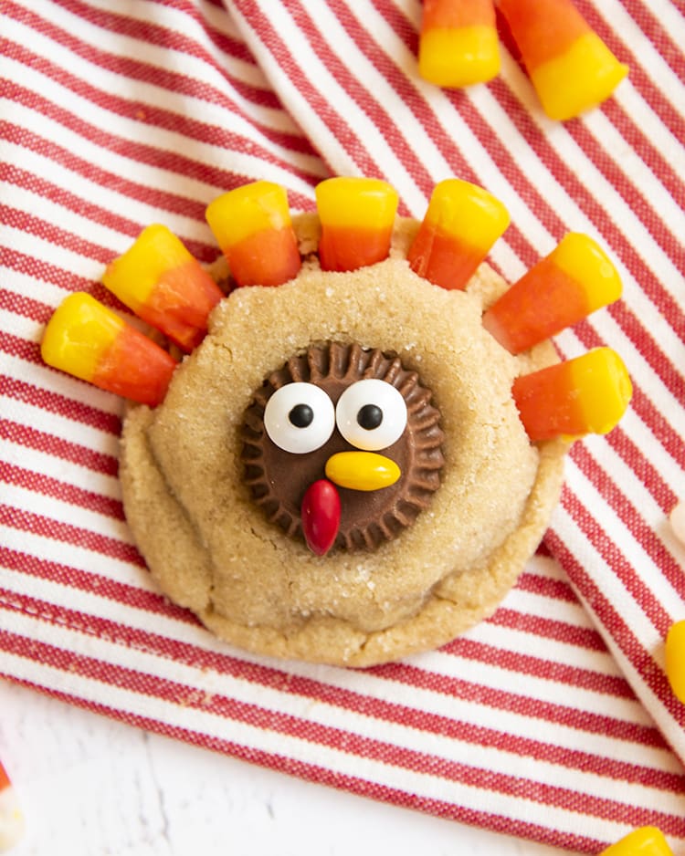 A close up shot of a peanut butter cookie decorated to look like a turkey with a peanut butter cup decorated with candy eyes, and chocolate covered candies for a beak, and candy corn at the top for feathers.