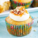 A carrot cake cupcake in a spring cupcake liner, topped with cream cheese frosting, that is topped with walnuts, and shredded carrots.