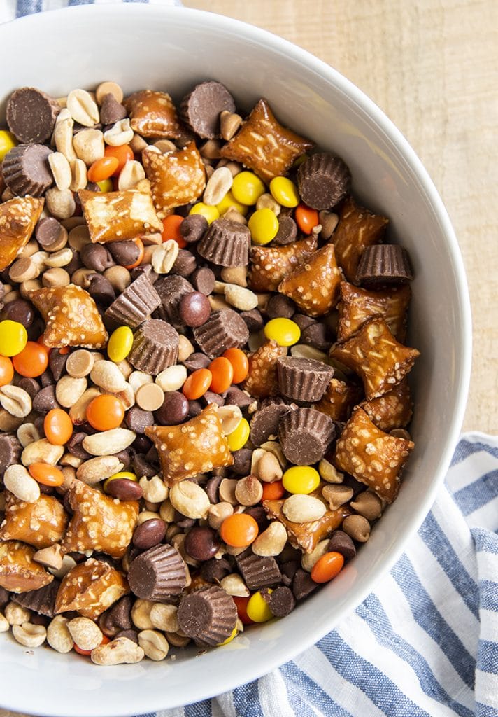 An overhead shot of a white bowl full of a Reeses Snack Mix, it has pretzels, peanuts, chocolate chips, peanut butter chips, reeses pieces, and mini reeses cups.