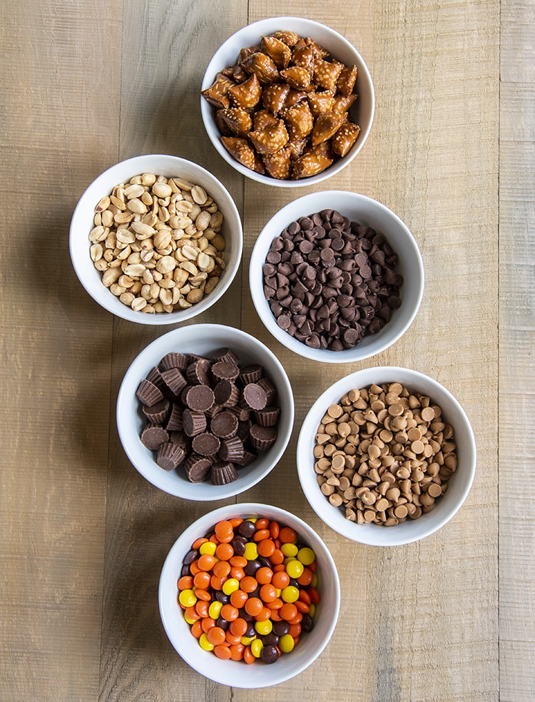 Small bowls of peanut butter filled pretzels, salted peanuts, chocolate chips, mini peanut butter cups, peanut butter baking chips, and reeses pieces.
