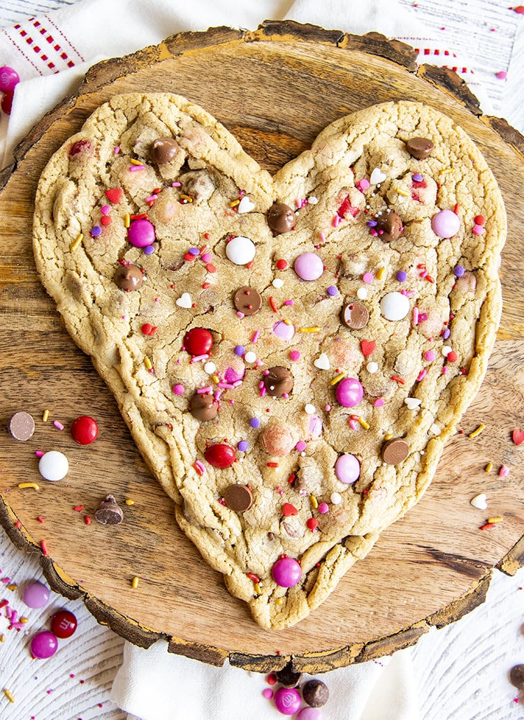 A big heart shaped cookie with valentine colored m&ms, chocolate chips, and valentine sprinkles set on a round wooden platter.