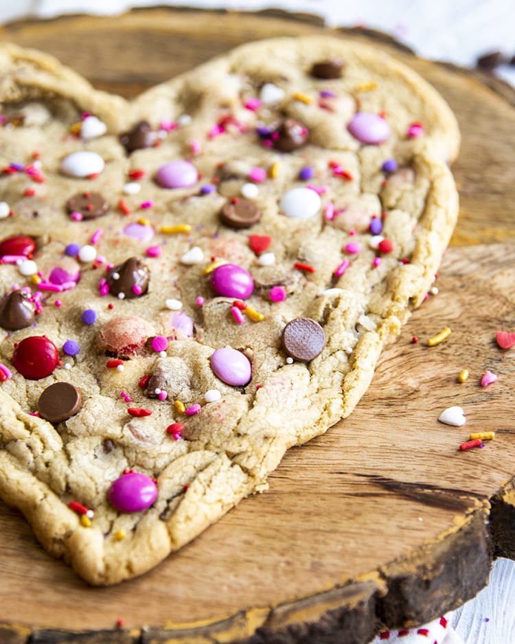 A heart shaped cookie on a wooden board with pink m&ms on top.