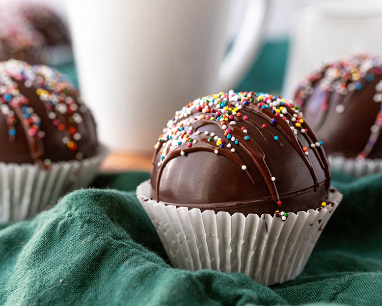 A close up of a chocolate dome ball, presumably a hot chocolate bomb, drizzled with chocolate and covered in rainbow colored non paireils.