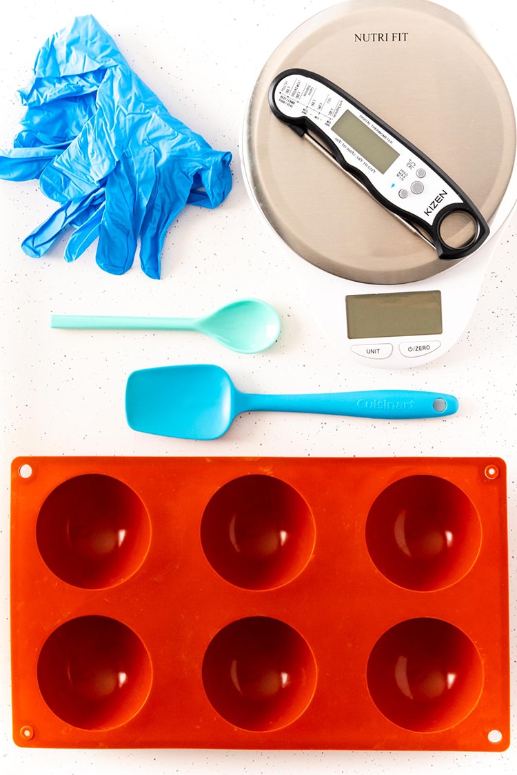 A photo showing rubber gloves, a food scale, a food thermoter, a small spoon, rubber spatula, and hot chocolate bomb silicone mold.