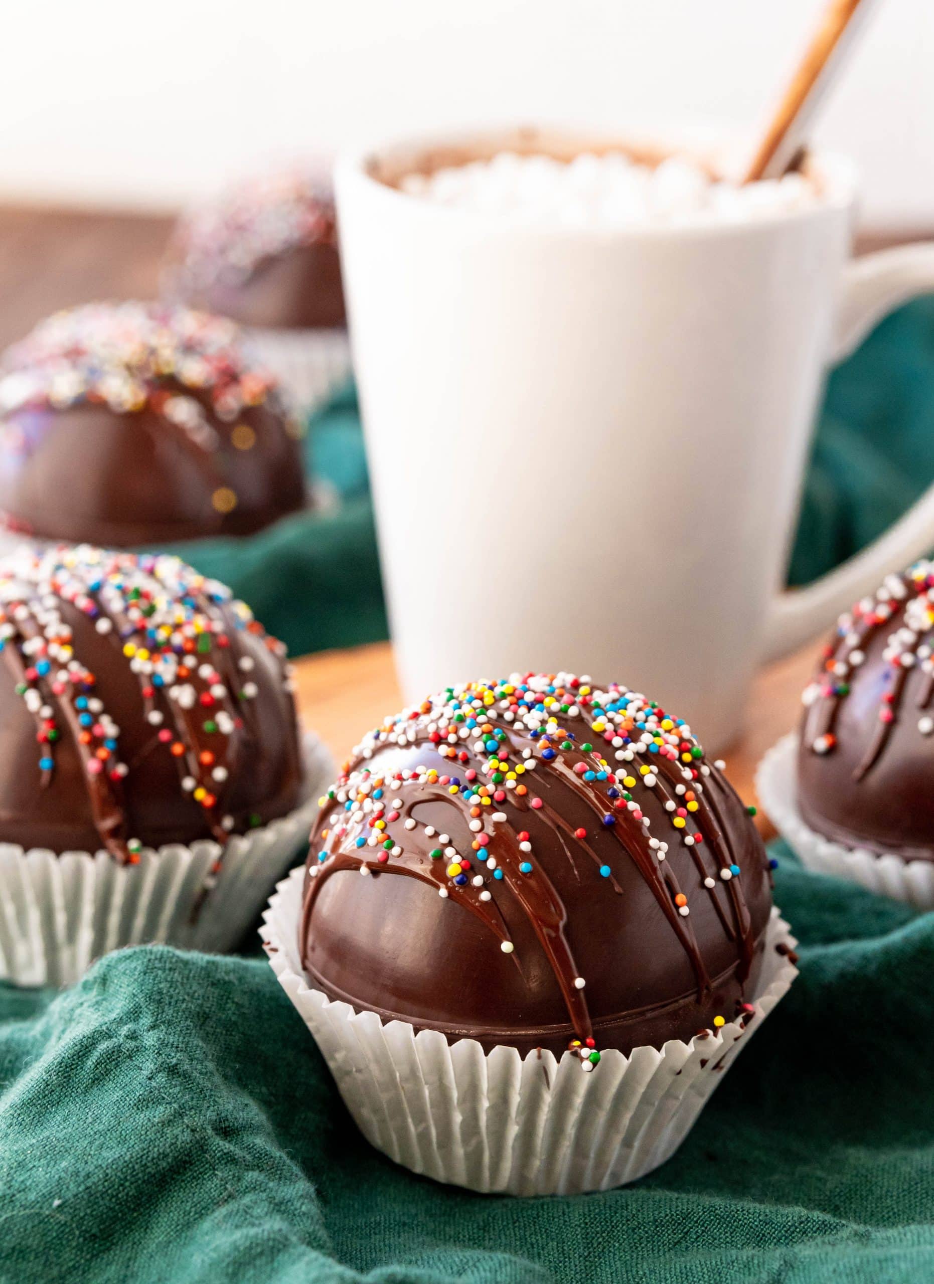 A hot chocolate bomb decorated in sprinkles with a mug of hot chocolate behind it.