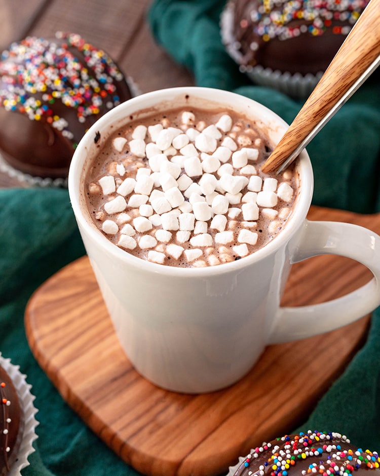 A white mug full of hot chocolate and mini marshmallows, with a wooden spoon in it.