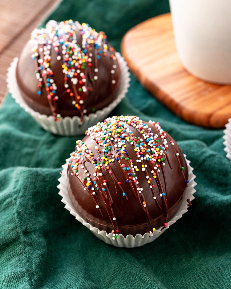 A close up of a chocolate dome ball, presumably a hot chocolate bomb, drizzled with chocolate and covered in rainbow colored non paireils.