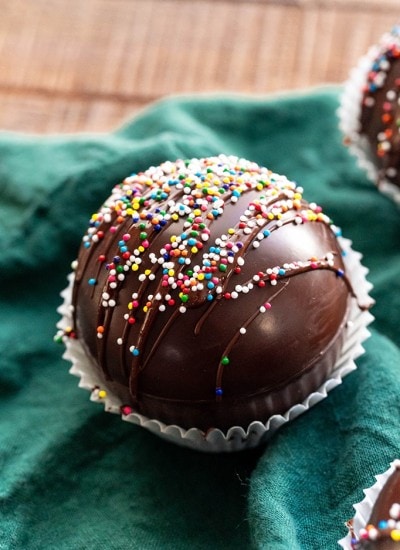A close up of a chocolate dome ball, presumably a hot chocolate bomb, drizzled with chocolate and covered in rainbow colored non paireils.