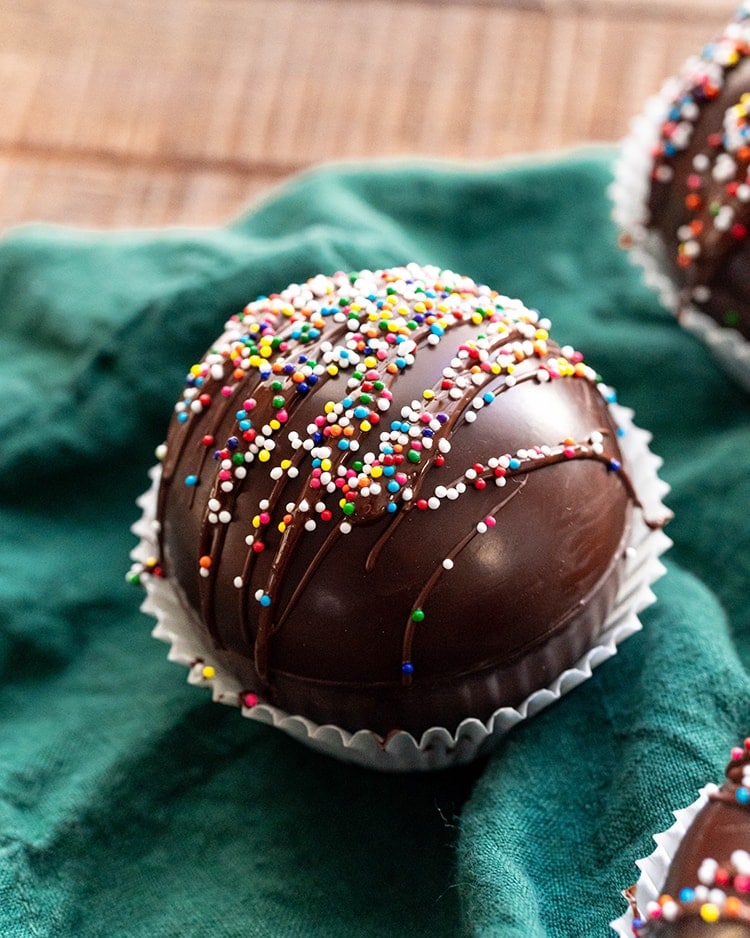 A close up of a chocolate dome ball, presumably a hot chocolate bomb, drizzled with chocolate and covered in rainbow colored non paireils.