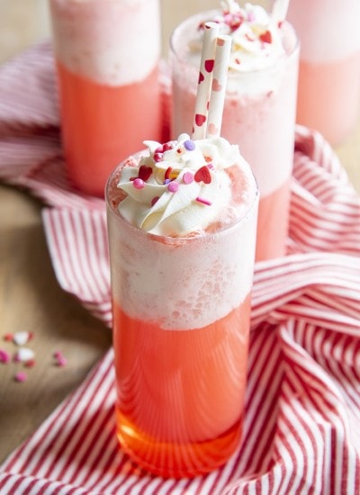 A tall glass cup filled with a pink drink, with foam on the top, and topped with whipped cream.