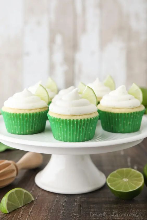Cupcakes on a cake stand with green cupcake liners, and piled with a white frosting on top, and a lime slice on each.