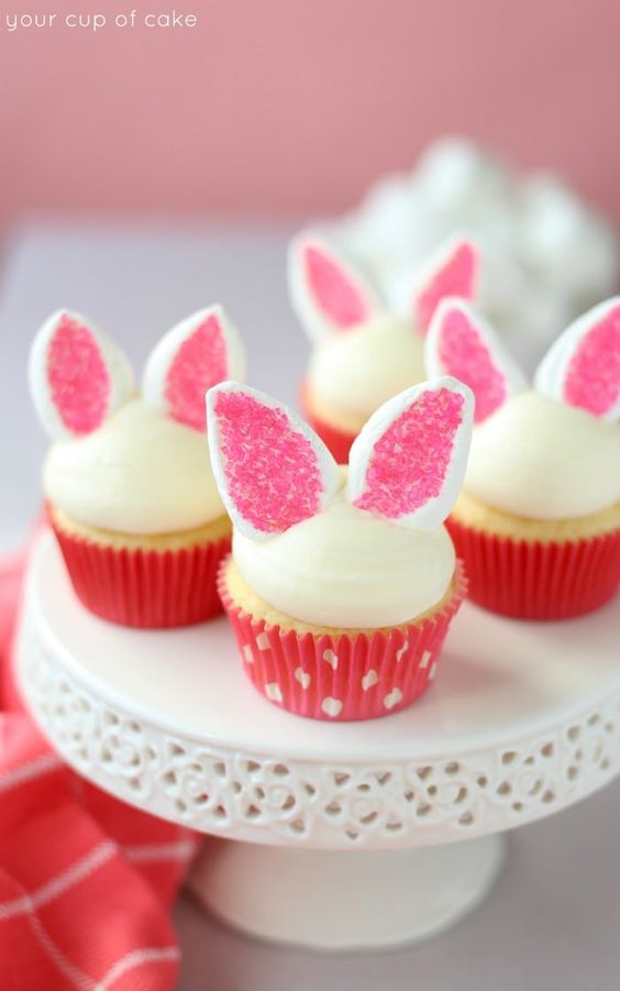 Four cupcakes on a cake stand with a half dome of white frosting and marshmallows cut in half and dipped in pink sprinkles to look like bunny ears on them.