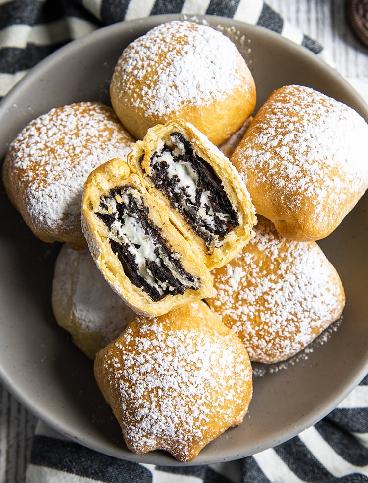 Air Fried Oreos in a bowl covered in powdered sugar, and there is one on top that is cut in half showing the Oreo in the middle.