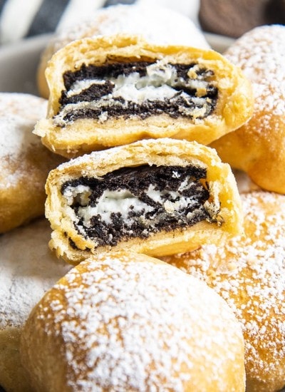 Air Fried Oreos in a bowl covered in powdered sugar, and there is one on top that is cut in half showing the Oreo in the middle.