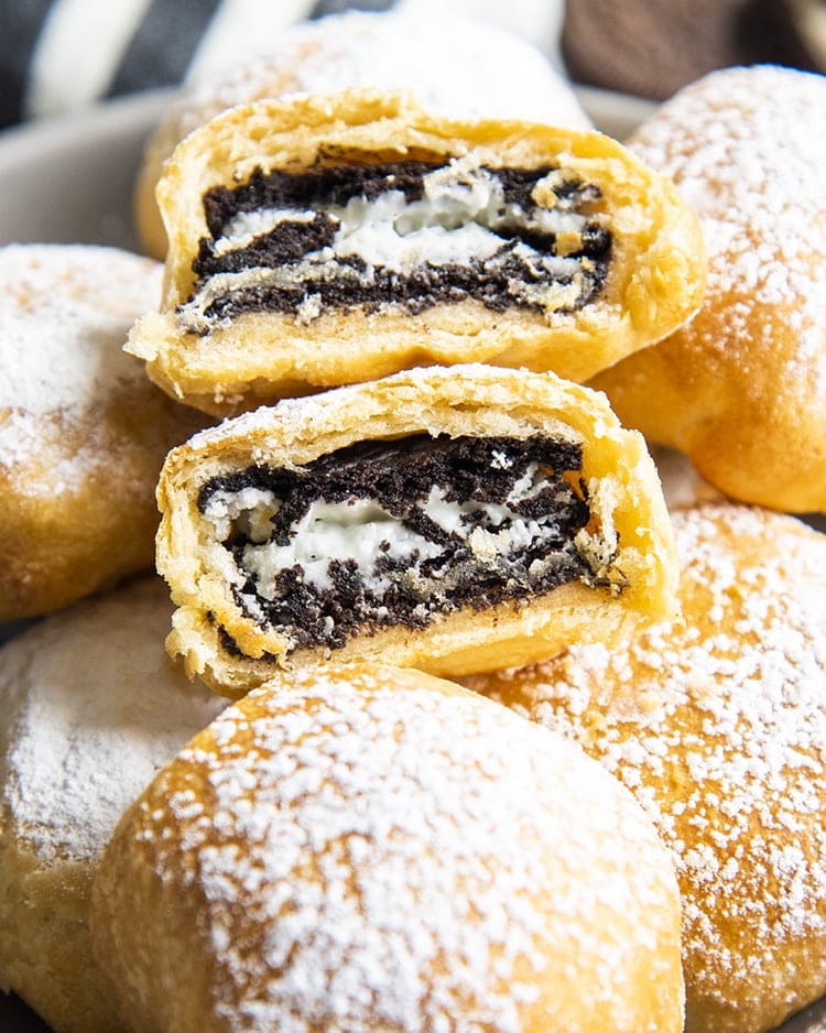 Air Fried Oreos in a bowl covered in powdered sugar, and there is one on top that is cut in half showing the Oreo in the middle.