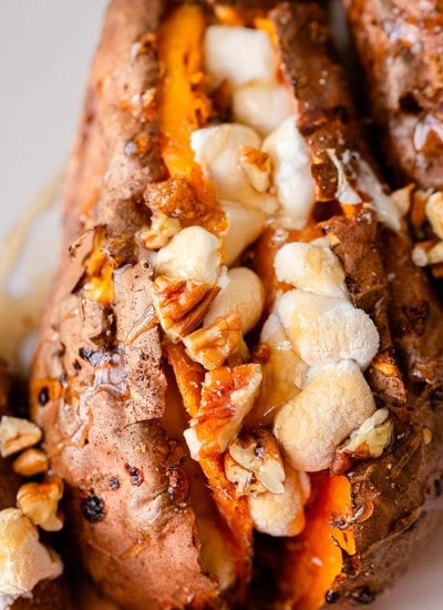 A close up of a baked sweet potato full of brown sugar, toasted marshmallows and pecans, with maple syrup drizzled over the top.