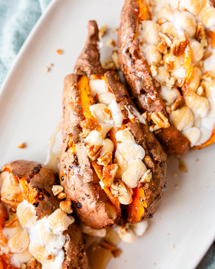 A row of three sweet potatoes loaded with toasted marshmallows and chopped pecans on a white plate.