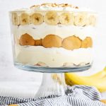 A layered banana pudding trifle in a trifle dish, with layers of Nilla Wafers, Banana Slices, Vanilla Pudding, and Whipped Cream.