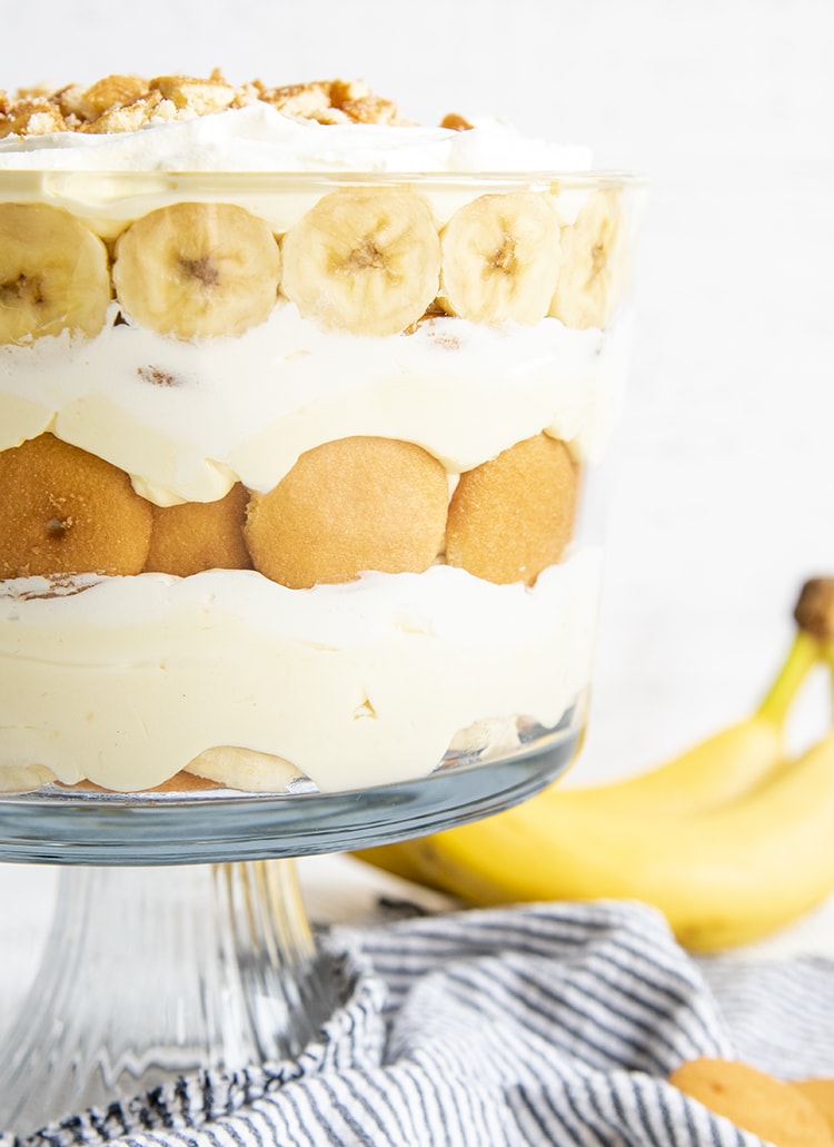 A close up of a layered banana pudding trifle in a trifle dish, with layers of Nilla Wafers, Banana Slices, Vanilla Pudding, and Whipped Cream.