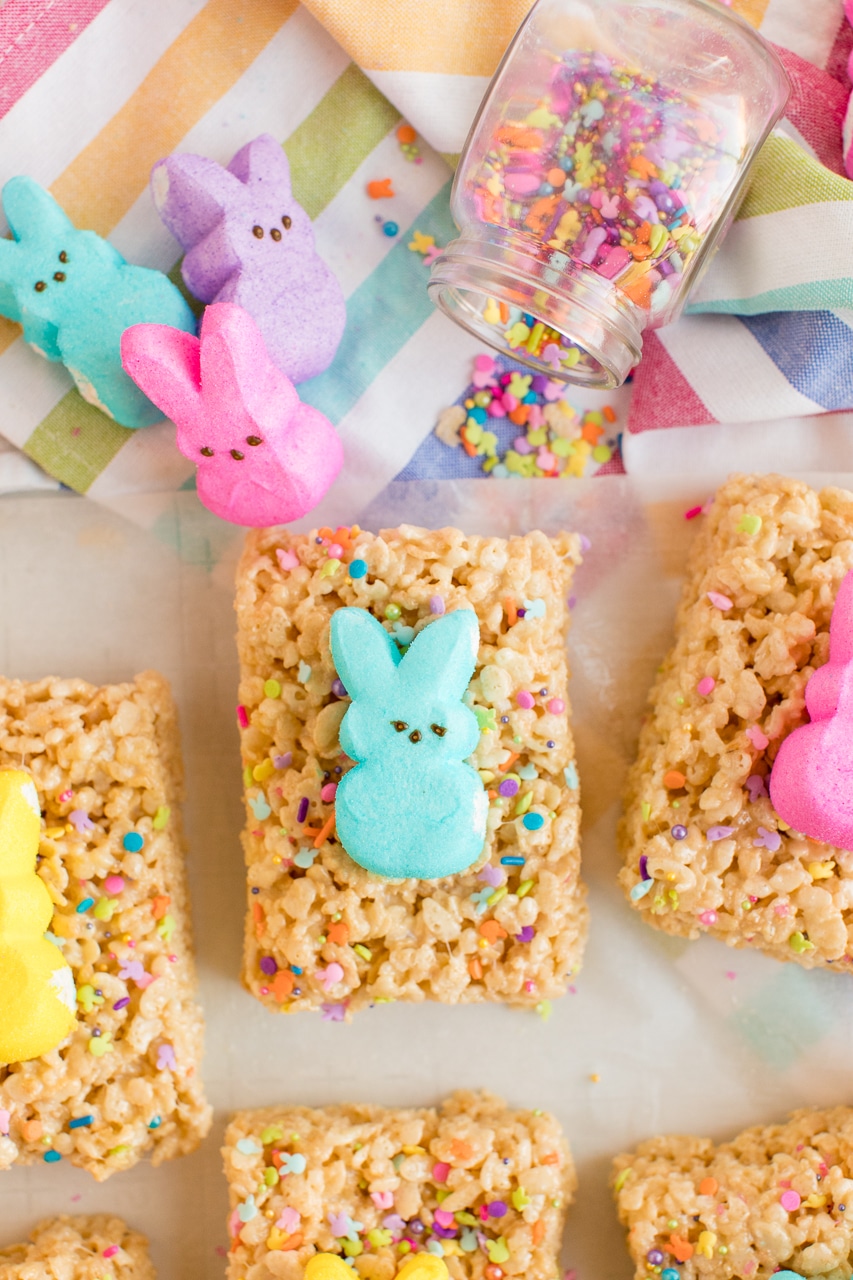 Rice Krispie treats decorated with funfetti colored sprinkles and topped with a Peeps bunny. There are more peeps laying along side them.