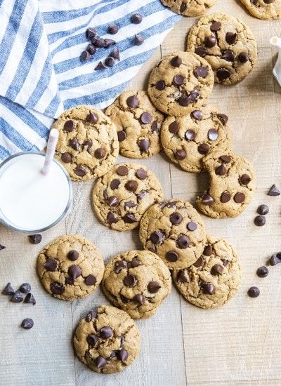 Flourless peanut butter chocolate chip cookies arranged randomly, slightly piled on a wooden surface, with a blue and white stripe cloth next to them. There is a glass of milk with a straw in it next to the cookies.