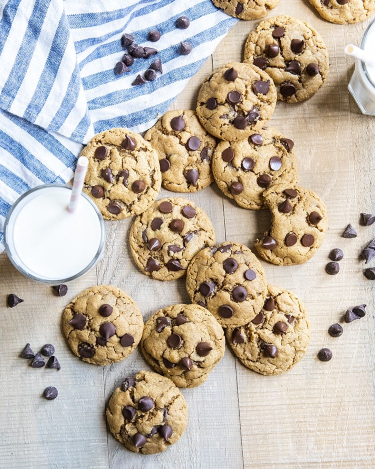 Flourless peanut butter chocolate chip cookies arranged randomly, slightly piled on a wooden surface, with a blue and white stripe cloth next to them. There is a glass of milk with a straw in it next to the cookies.