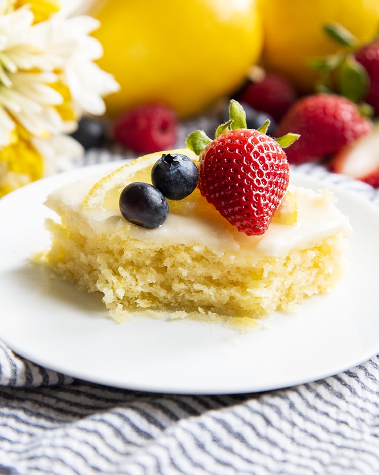 A slice of a yellow sheet cake on a plate that is topped with two blueberries, a strawberry, and lemon slice with a bite out of the front.