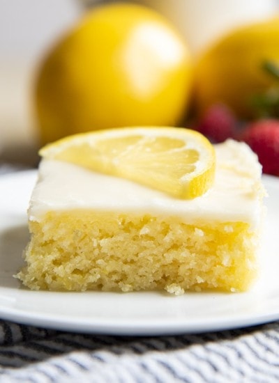 A close up of a slice of lemon sheet cake topped with a white icing, and a half slice of a lemon.