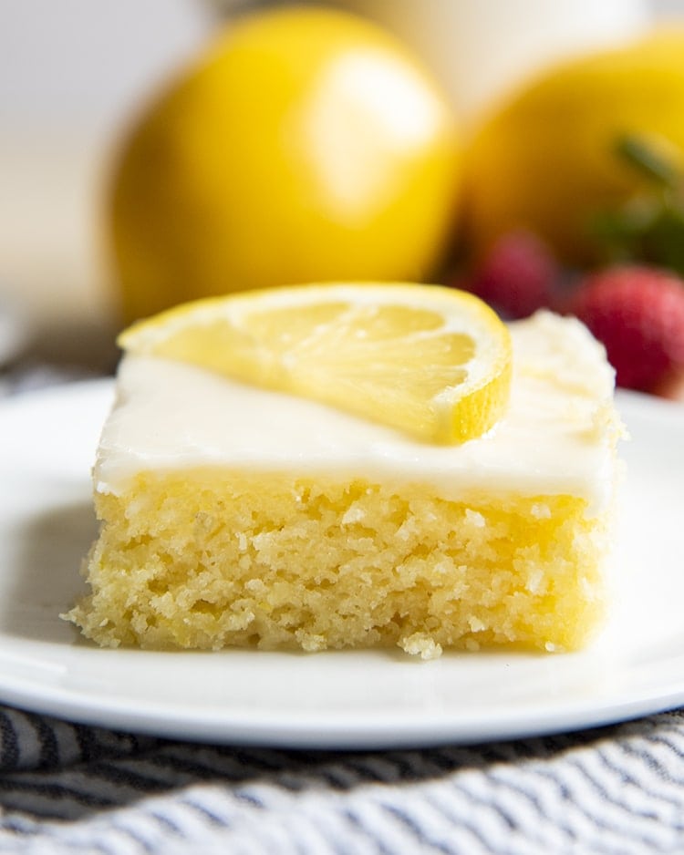A close up of a slice of lemon sheet cake topped with a white icing, and a half slice of a lemon.