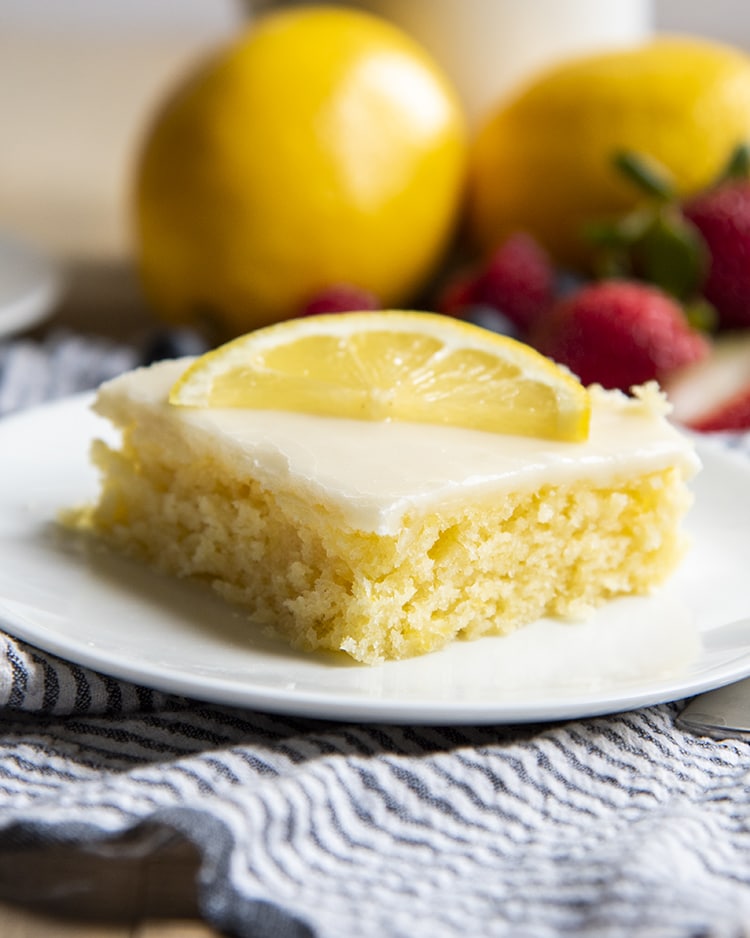 A close up of a slice of lemon sheet cake topped with a white icing, and a half slice of a lemon.