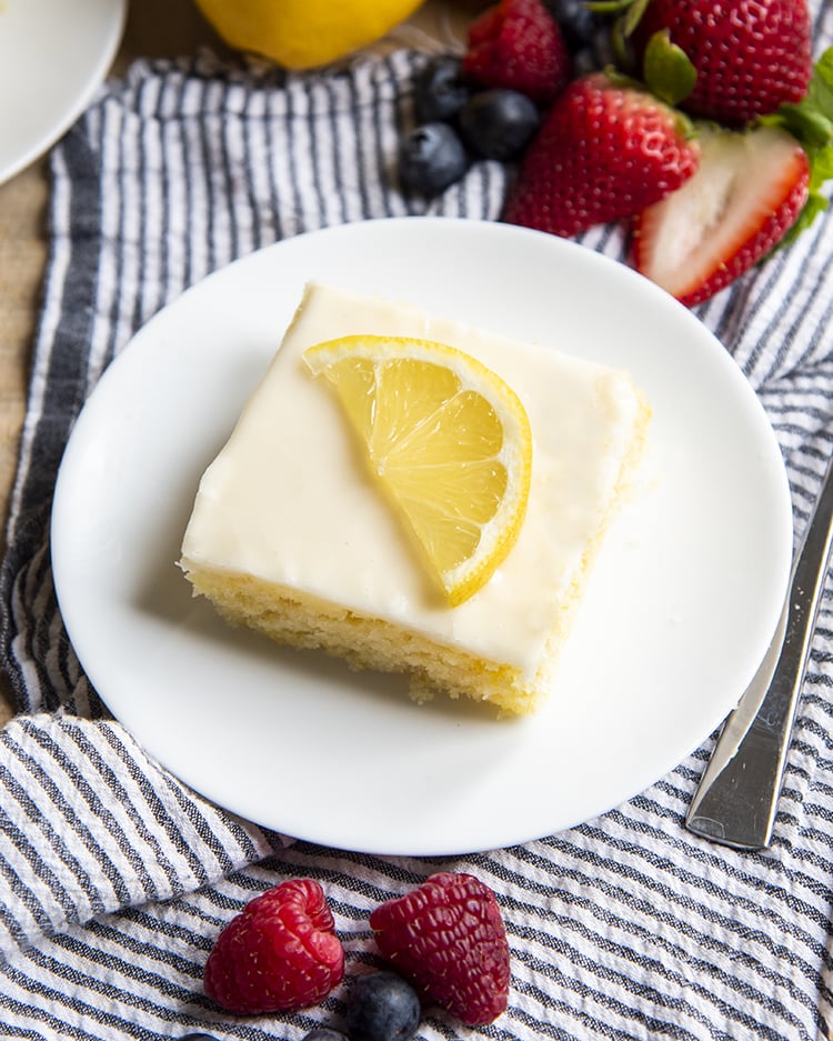 An overhead shot of a slice of lemon sheet cake on a white plate topped with a lemon slice.