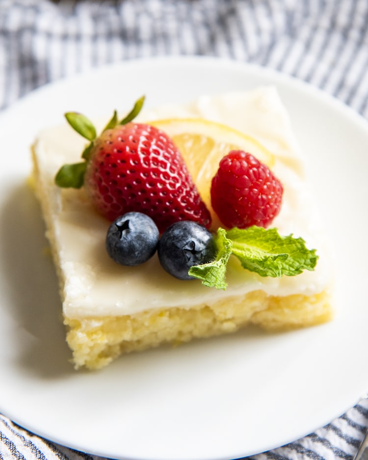 A slice of lemon sheet cake on a plate topped with fresh berries and mint leaves.