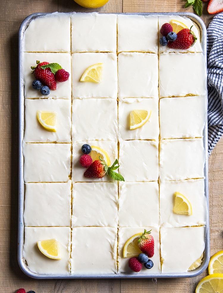 A baking pan full of cake shot from the top showing the icing, and cake cut into slices. Some of the slices have a quarter of a lemon slice and some berries.
