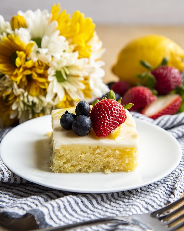A slice of lemon sheet cake on a plate topped with fresh berries, and there are flowers behind the plate.