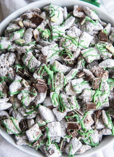 An overhead shot of mint muddy buddies in a white bowl. They are drizzled with green candy melts and there are chopped up Andes Mint pieces mixed in.