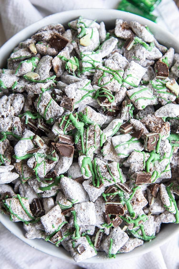 An overhead shot of mint muddy buddies in a white bowl. They are drizzled with green candy melts and there are chopped up Andes Mint pieces mixed in.