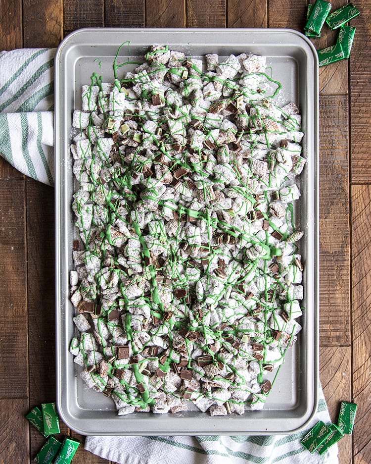 A baking pan covered in chex muddy buddies covered in powdered sugar. There are small pieces of Andes mints spread around, and green candy melts drizzled across the top.