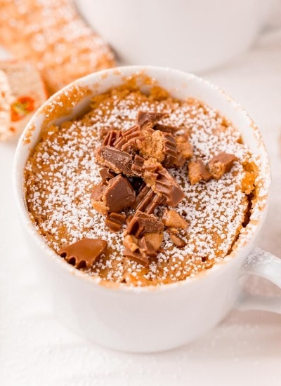 A nutter butter mug cake in a white mug sprinkled with powdered sugar, and topped with chopped peanut butter cups.