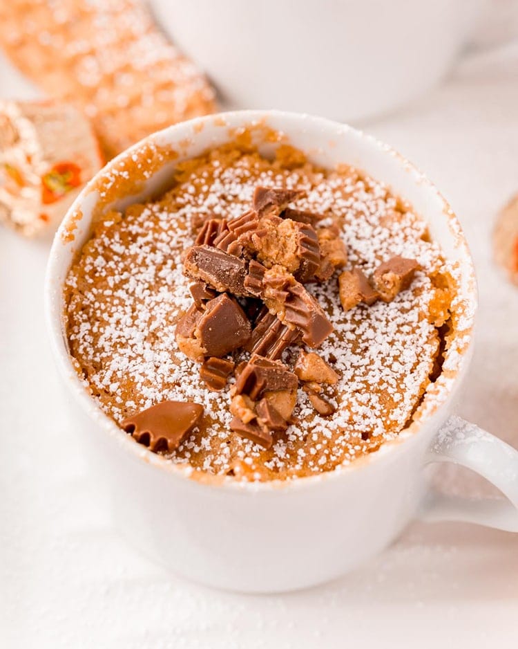 A nutter butter mug cake in a white mug sprinkled with powdered sugar, and topped with chopped peanut butter cups.