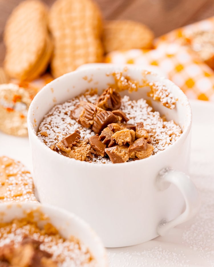 A nutter butter mug cake in a white mug sprinkled with powdered sugar, and topped with chopped peanut butter cups.
