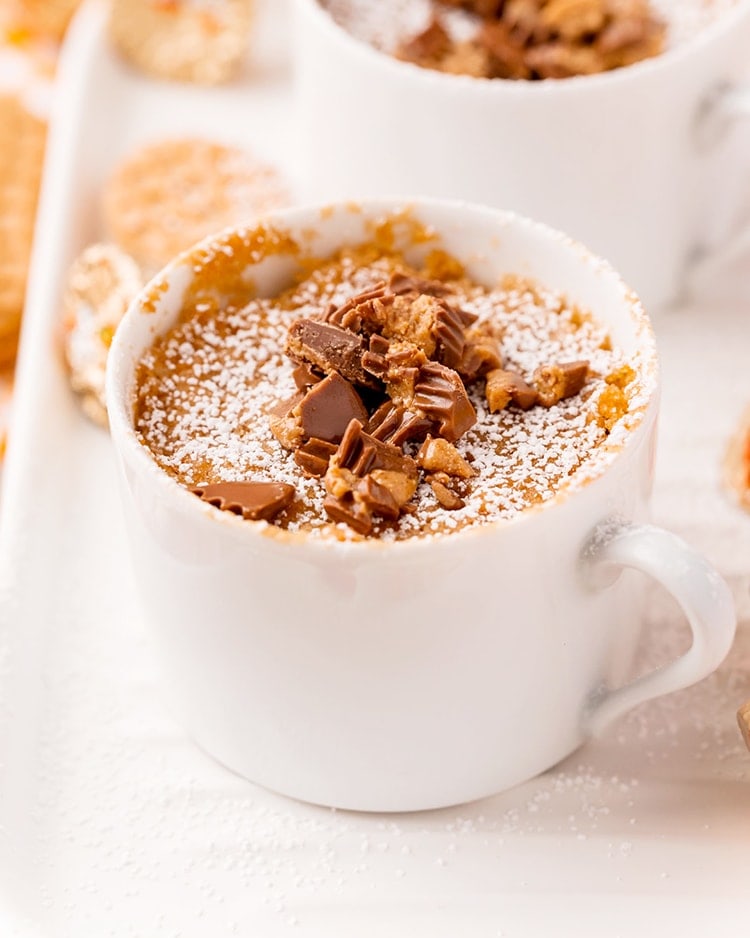 A nutter butter mug cake in a white mug sprinkled with powdered sugar, and topped with chopped peanut butter cups.