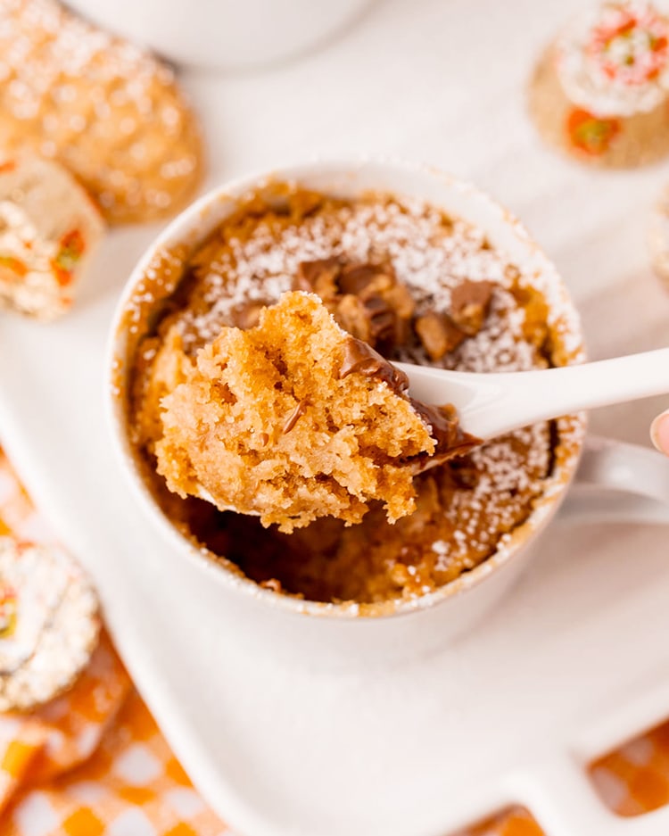 A spoon holding a bite out of a peanut butter mug cake, it looks cakey, and orangeish colored.