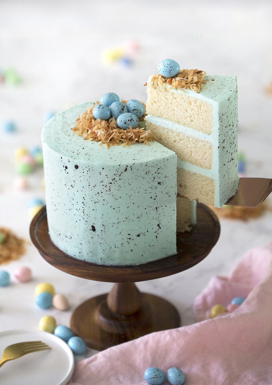 A three layered white cake, with a blue speckled frosting, topped with toasted coconut and blue candied eggs on top.