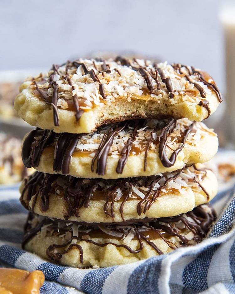 A stack of 5 caramel, coconut sugar cookies drizzled with chocolate over the top. The top cookie has a bite out of it showing the soft cookie, and the cookie has chocolate on the bottom.