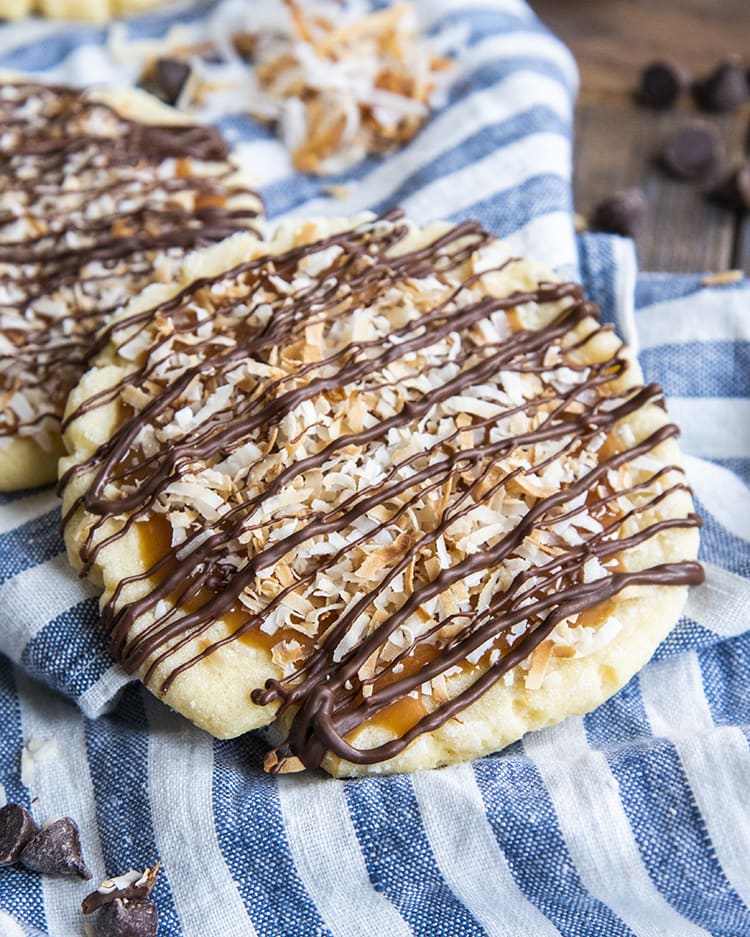 A sugar cookie topped with caramel, toasted coconut, and drizzled with chocolate, leaning on another cookie on a blue and white cloth.