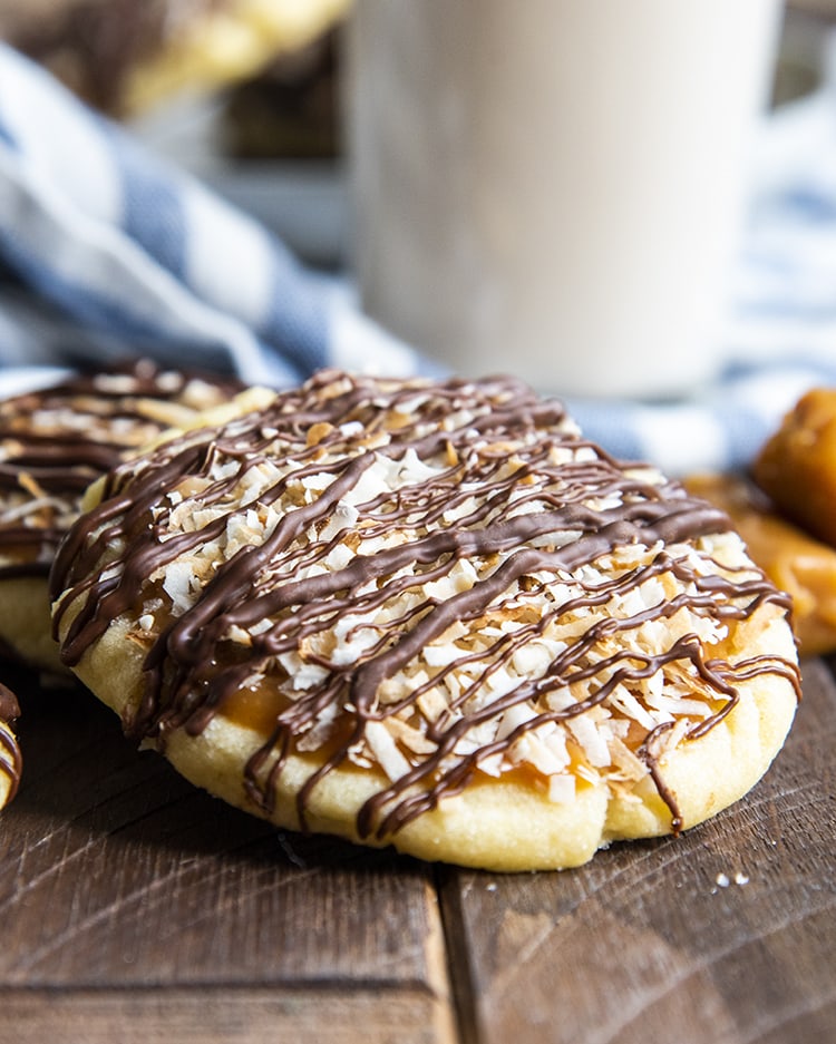 A sugar cookie topped with caramel, toasted coconut, and drizzled with chocolate, leaning on another cookie.